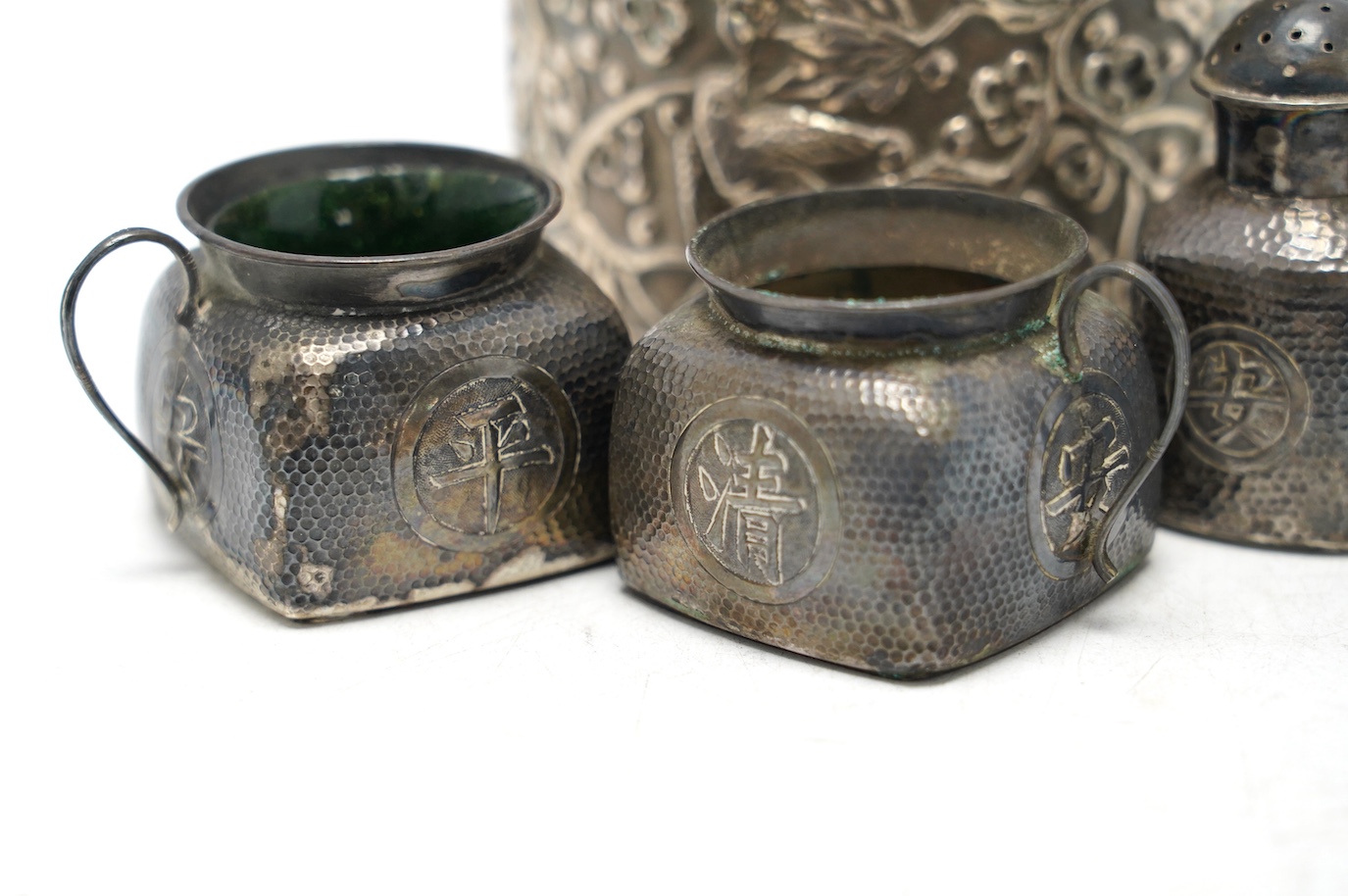 A Chinese embossed white metal cylindrical jar and cover, 11.2cm, and a similar white metal four piece cruet set. Condition - poor to fair